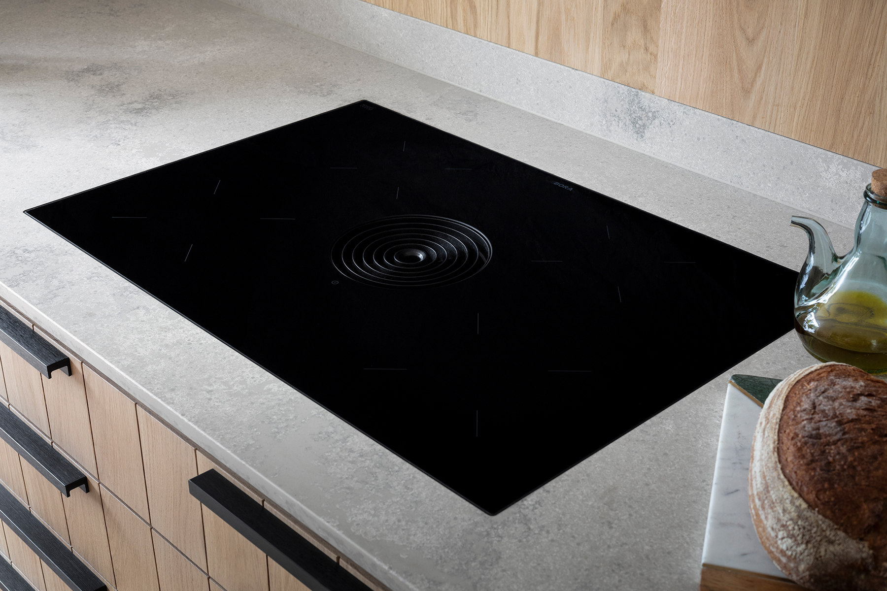 A modern kitchen countertop with a sleek black induction cooktop integrated into the light-colored stone surface. The cooktop features a central circular vent and subtle markings indicating burner locations. On the right side of the countertop, there is a glass bottle with a spout containing olive oil and a loaf of bread on a cutting board.