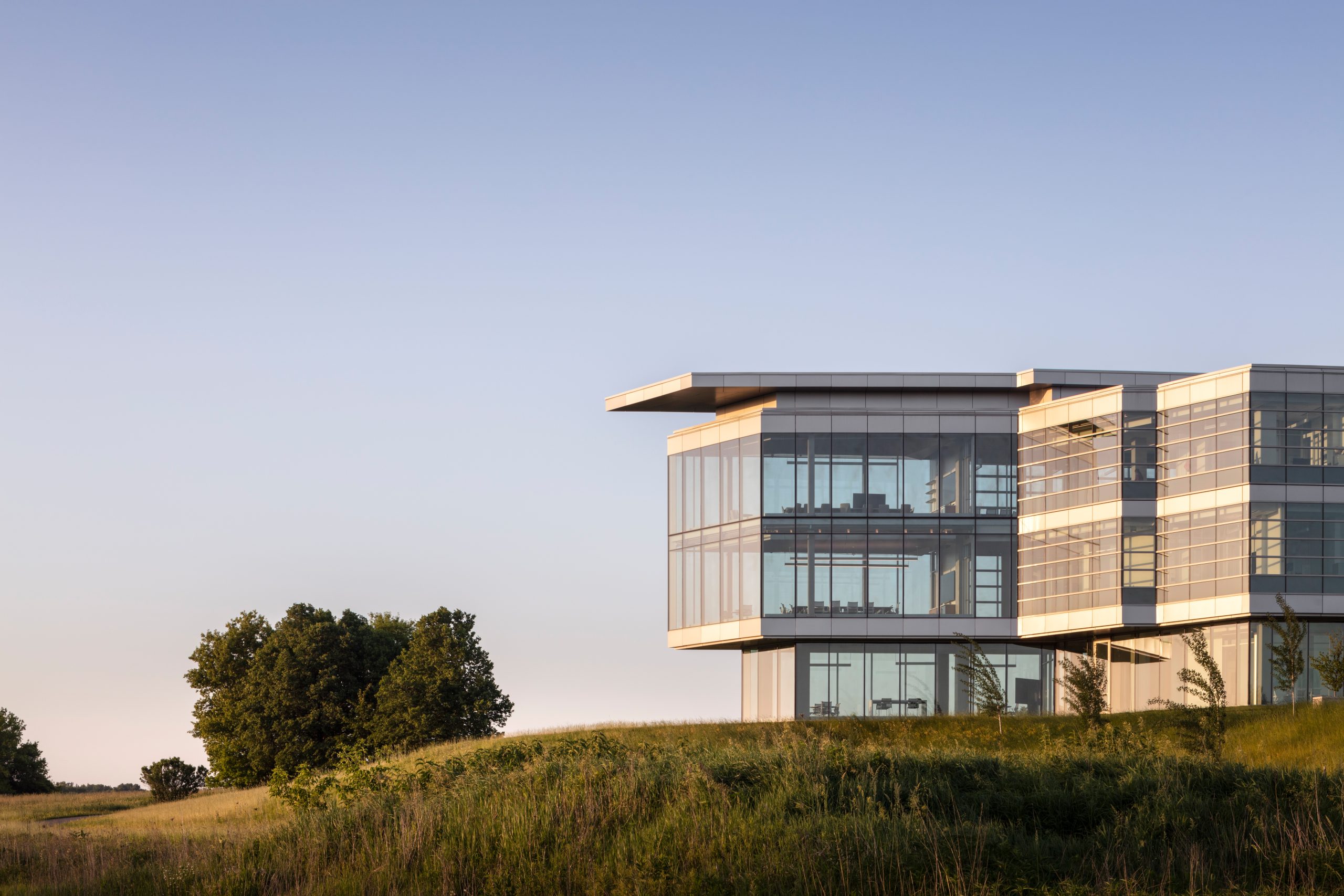 A modern glass building with a sleek, angular design is situated on a grassy hill. The building features large windows that reflect the surrounding landscape, including a cluster of trees to the left. The sky is clear with a gradient from light blue at the horizon to a deeper blue at the top. The image captures the building in natural light, highlighting its contemporary architecture and integration with the natural environment.