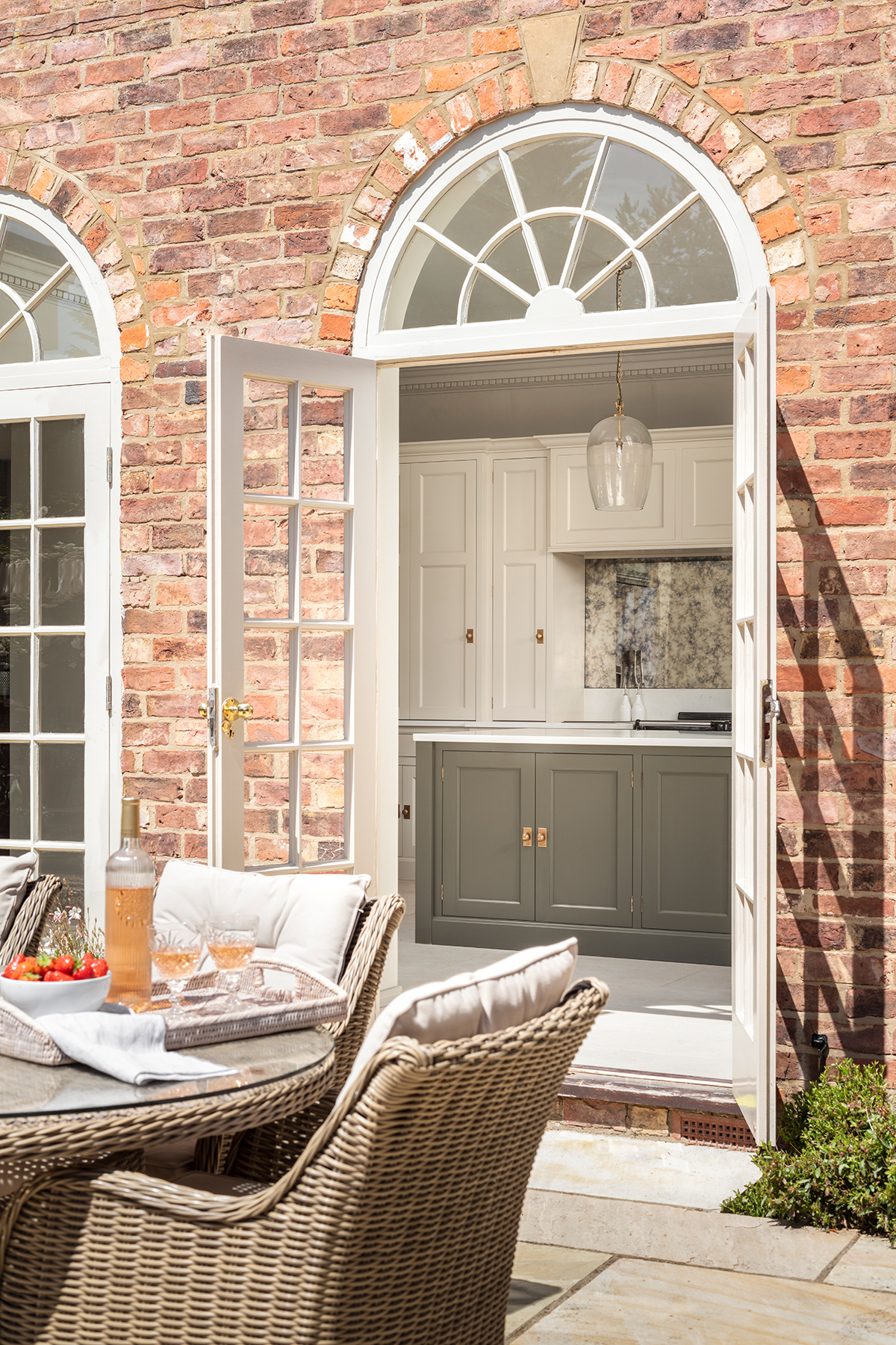 A patio with wicker furniture leading to a kitchen with gray and cream shaker cabinets through arched double doors with glass panels, set against a brick wall.