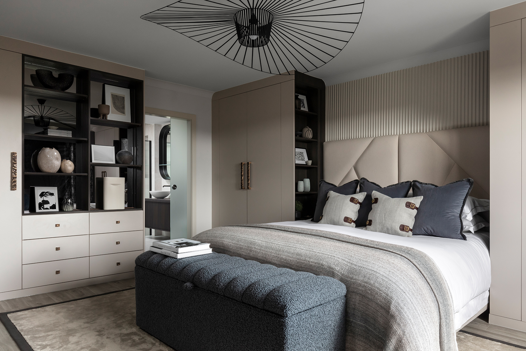 A well-designed bedroom with a modern aesthetic. The room features built-in wardrobes and storage, a large upholstered headboard, a comfortable bed with stylish pillows and a throw, and an intricate black ceiling fixture. An en-suite bathroom is visible through a doorway.