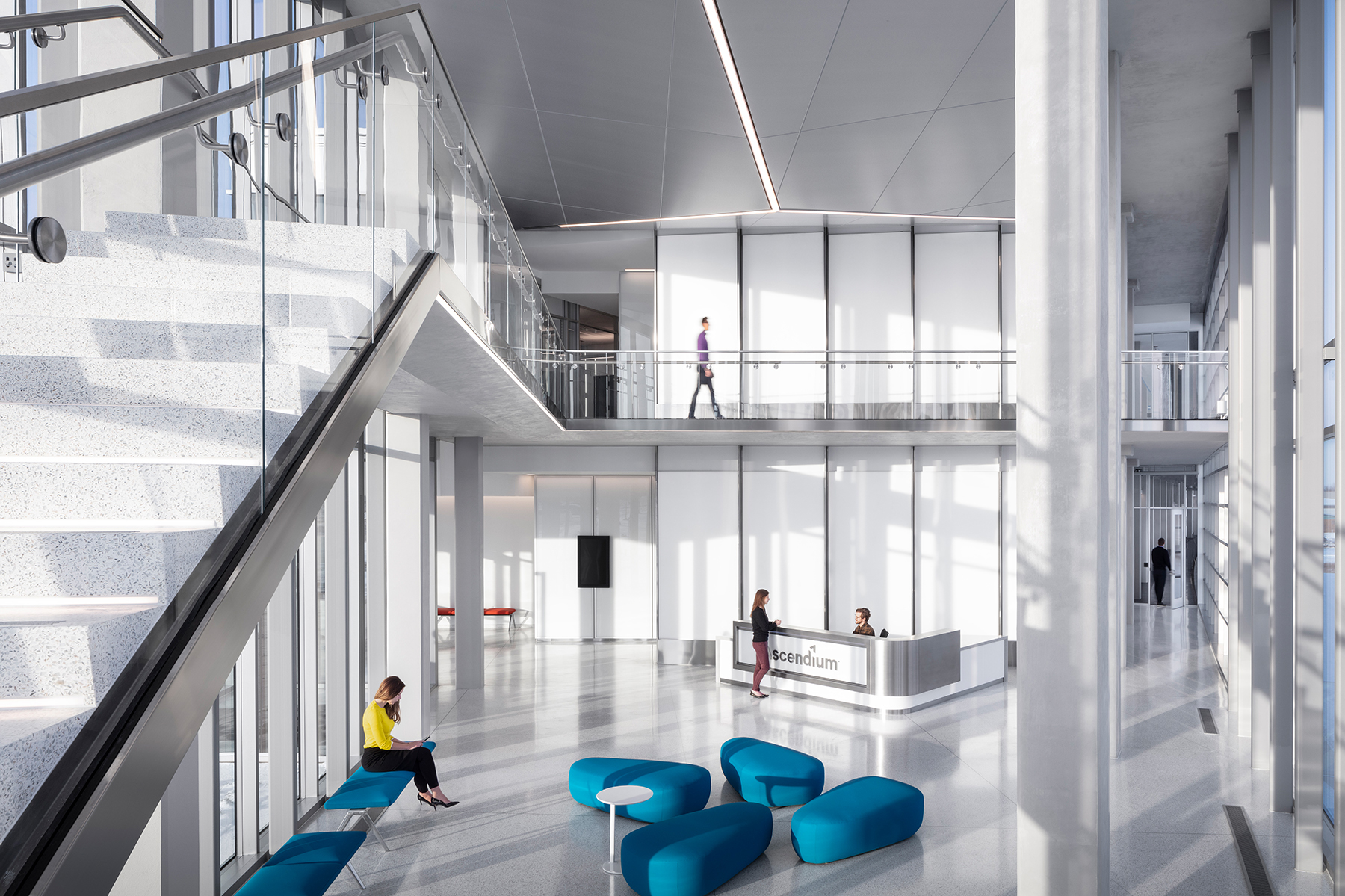 A modern, spacious office lobby with large windows allowing natural light to flood the area. The lobby features a sleek staircase with glass railings on the left, leading to an upper floor where a person is walking. In the center, there are several blue, uniquely shaped seating arrangements and a small white table. A person in a yellow top is seated on one of the benches, looking at a device. To the right, a reception desk with the name 'Ascendium' is staffed by a person, while another person stands in front of the desk. The overall design is clean and minimalist, with a focus on open space and light.