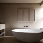 A bright and elegant bathroom with a light beige color scheme. An oval, freestanding white bathtub takes center stage, complemented by a modern vanity with a black faucet and stacked white towels. Three framed textured art pieces hang on the wall, and natural light streams in through a nearby window. A small, round table with a candle adds to the ambiance.