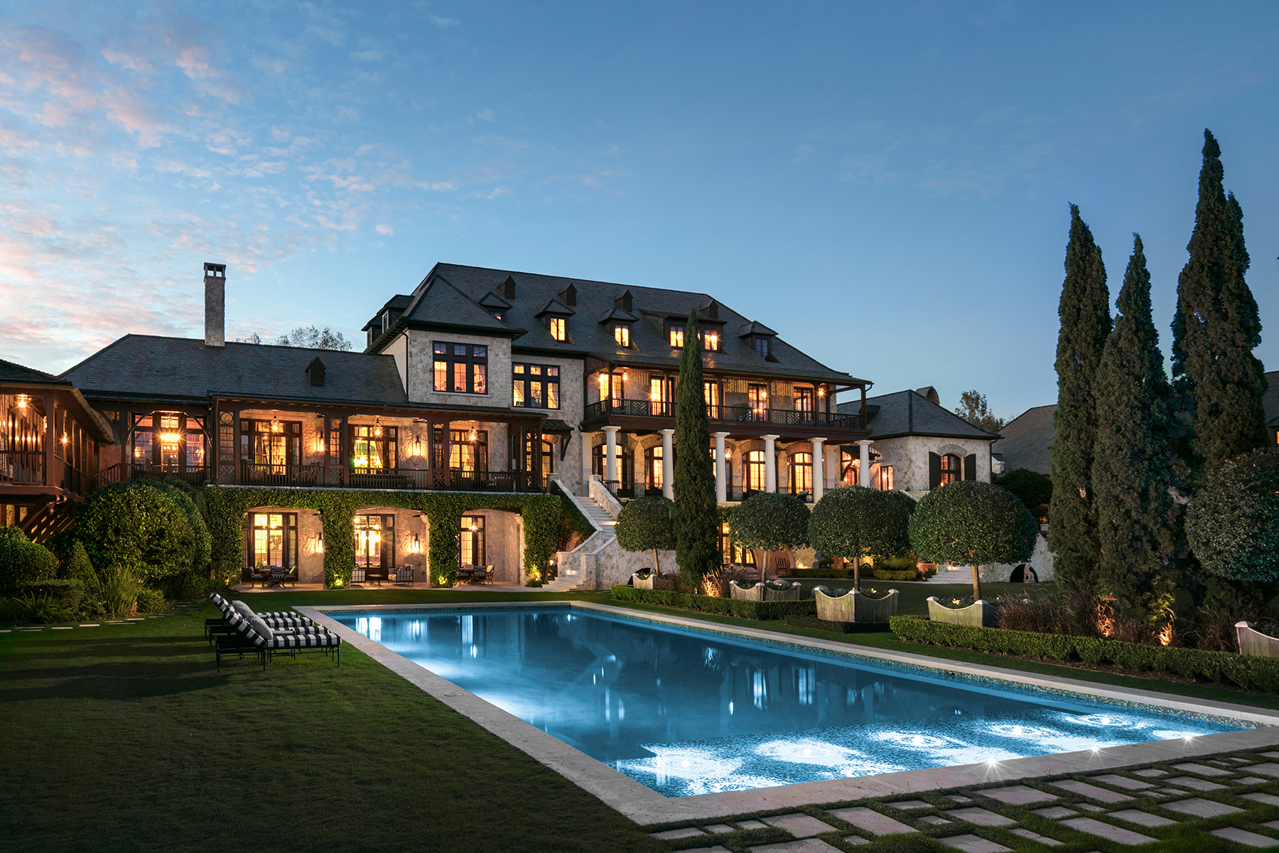 A stately home with a French chateau architectural influence, showcasing a symmetrical facade, multiple balconies with metal railings, a lit swimming pool, and well-defined landscaping against a dusk sky.