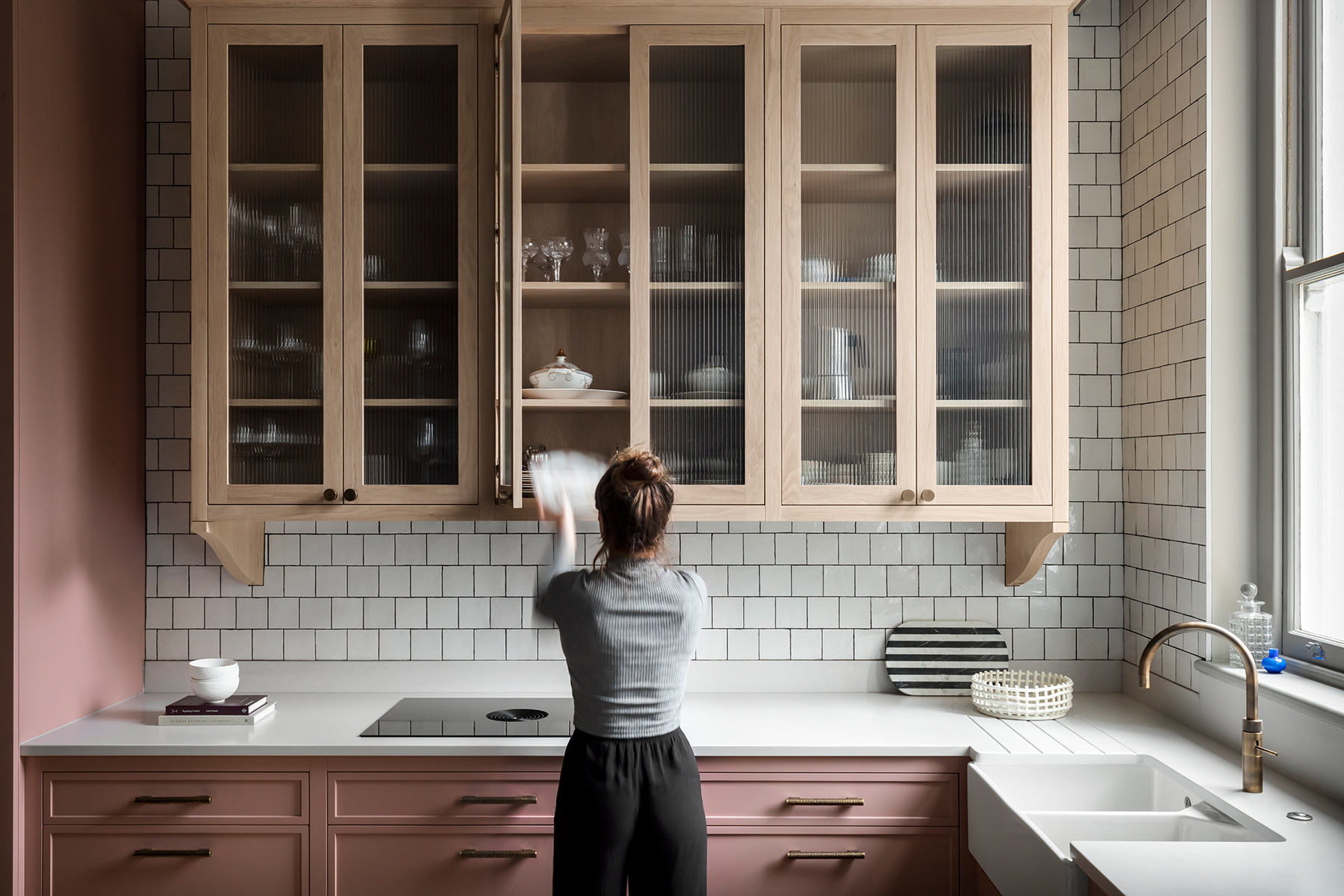Woman organizing glass cabinets above a pink kitchen counter.